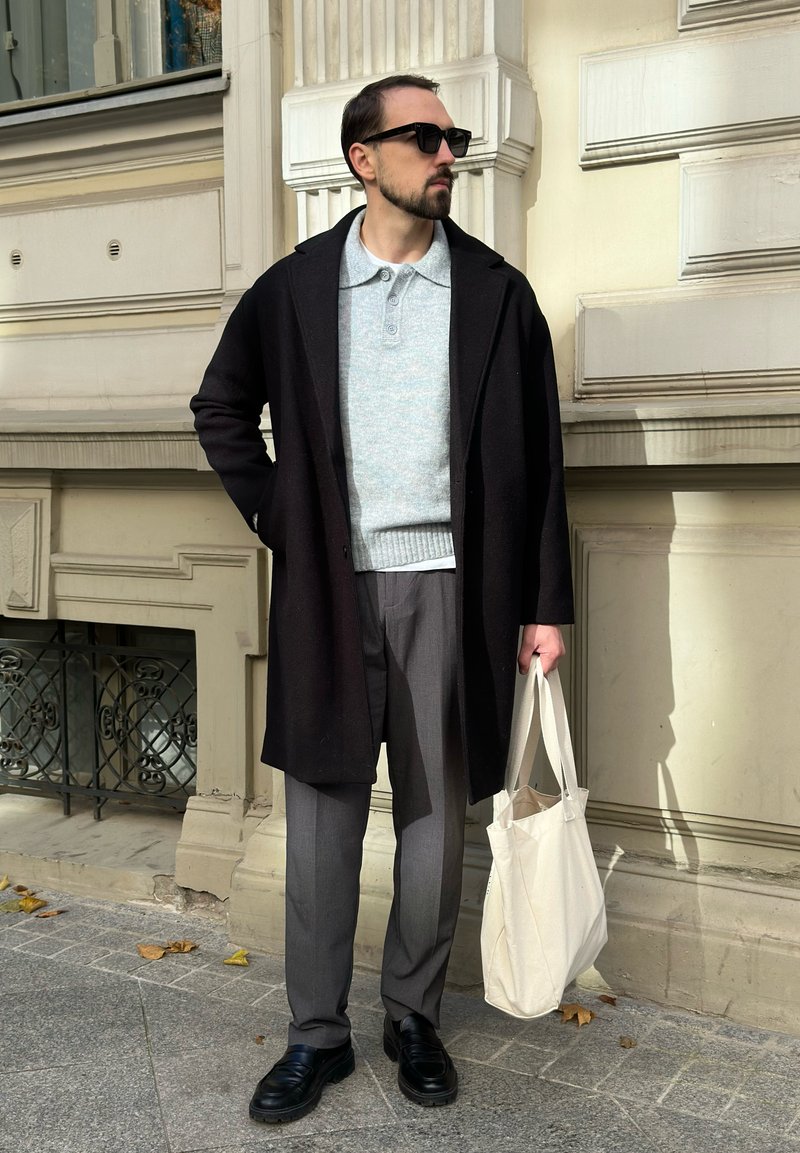 Mann in schwarzem Mantel, grauem Pullover, grauen Hosen, schwarzen Schuhen und Sonnenbrille, der eine weiße Tragetasche hält und auf dem Bürgersteig einer Stadt neben einem beigefarbenen Gebäude steht.