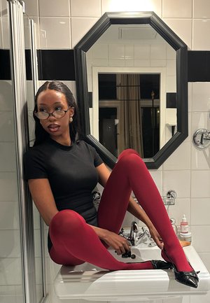 Mujer con gafas, vestida con blusa negra y medias rojas, sentada en el lavabo del baño ajustando un zapato negro frente a un espejo octogonal.
