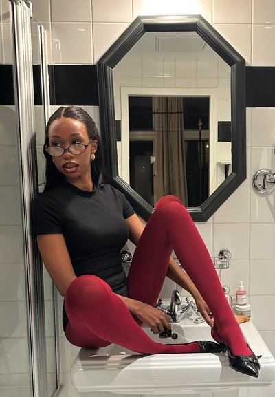 Mujer con gafas, vestida con blusa negra y medias rojas, sentada en el lavabo del baño ajustando un zapato negro frente a un espejo octogonal.