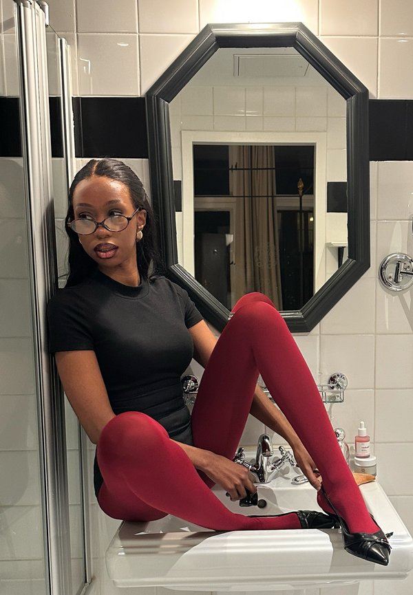 Femme portant des lunettes, un haut noir et des collants rouges, assise sur un lavabo de salle de bain, ajustant une chaussure noire devant un miroir octogonal.
