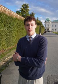 Giovane uomo in maglione blu navale e camicia con colletto, in piedi con le braccia incrociate, sorridente, in un giardino con un edificio storico sullo sfondo.