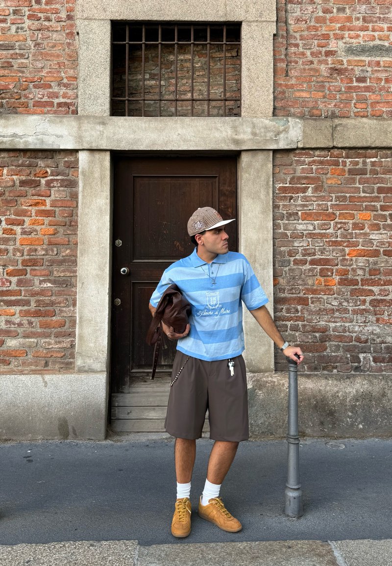 Polo shirt rayé bleu clair et blanc, shorts marron, chaussures marron, tenant un sac marron. Debout à côté d'un mur en pierre et d'une porte en bois.