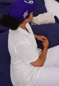 Vit stickad topp med korta ärmar, bärs över ljusfärgade byxor, i kombination med en lila basebollkeps. Sitter på mörka kuddar.