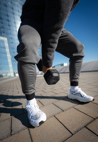 Melns hantelis, ko tur pelēkā flīsa un pieguļošās biksēs, kopā ar baltām sporta kurpēm. Virsma ir klāta ar taisnstūrveida akmeņiem.