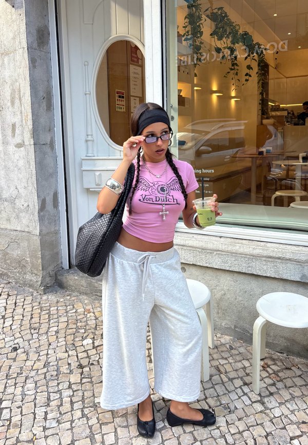 Jonge vrouw in een roze crop top en grijze wijde broek stelt haar zonnebril bij terwijl ze een groene drank vasthoudt buiten een café in een straat met kinderkopjes.