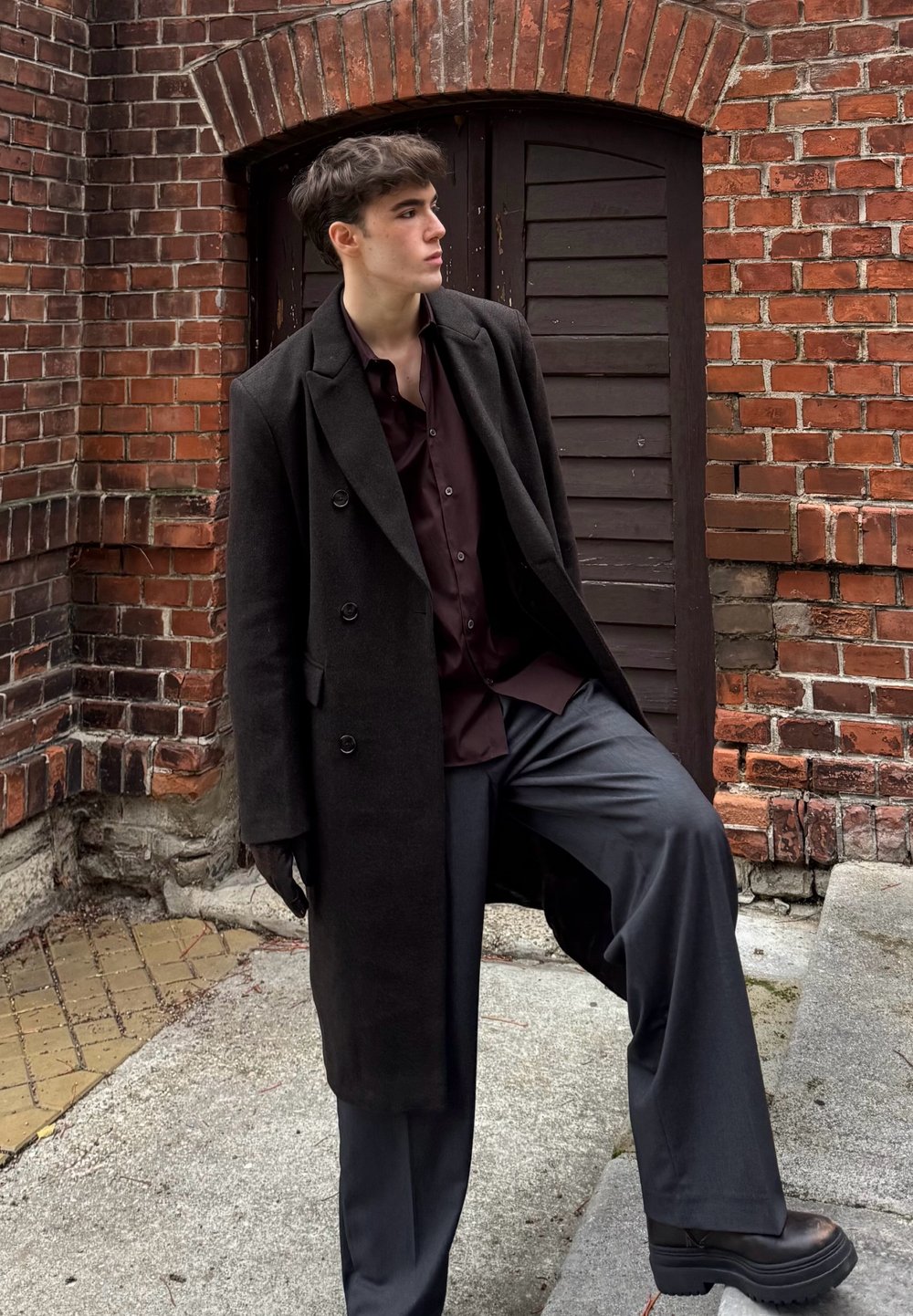 Sort dobbeltradet frakke over mørkerød skjorte med knapper, kombineret med mørkegrå bukser med vide ben og sorte platformstøvler, sat op mod en murstensvæg.