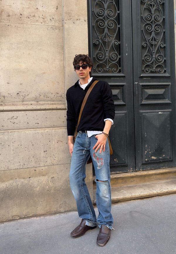 Junger Mann in schwarzem Pullover, zerrissenen Jeans mit Flicken, braunen Lederschuhen und Sonnenbrille, stehend vor einer Steinmauer und einer verzierten dunklen Tür.