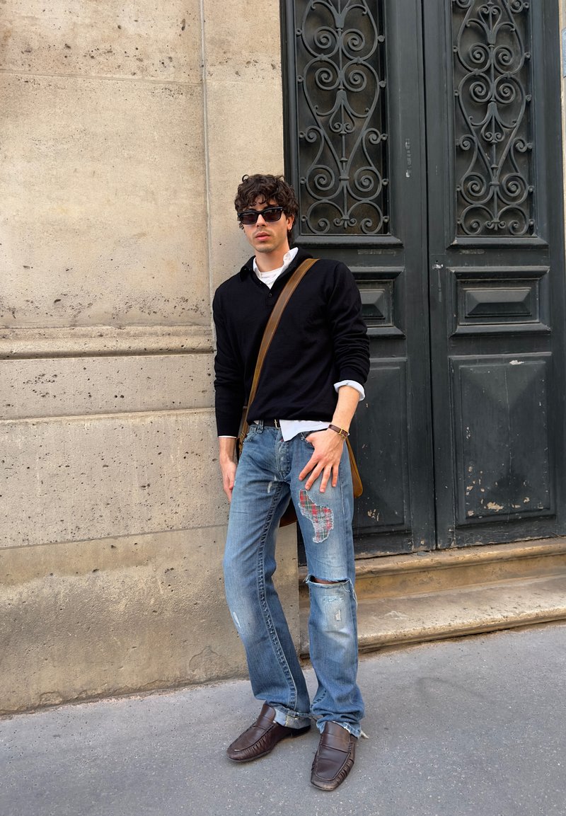 Young man wearing black sweater, patched ripped jeans, brown leather shoes, and sunglasses, standing against a stone wall and ornate dark door.