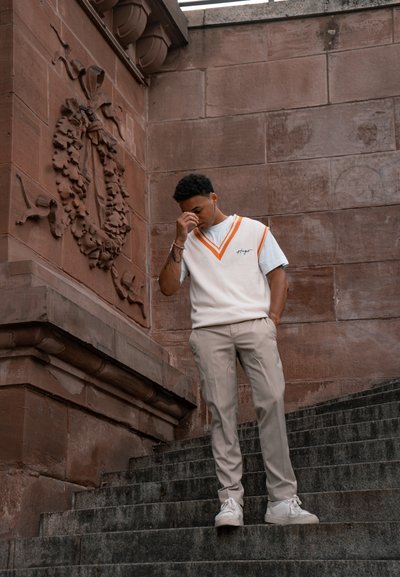 Chaleco de punto blanco con ribetes naranjas sobre una camisa azul, combinado con pantalones beige y zapatillas blancas, en unos escalones de piedra cerca de una pared decorativa.