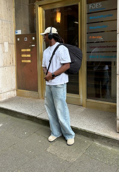 Mochila negra, jeans de corte holgado de color azul claro, camiseta blanca, zapatos beige, auriculares negros y una gorra beige clara, de pie junto a una puerta.