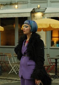 Costume sur mesure violet avec un manteau noir duveteux, cheveux bleu et violet, un maquillage des yeux bleu audacieux et de grandes boucles d'oreilles en argent. L'arrière-plan présente des sièges de café.