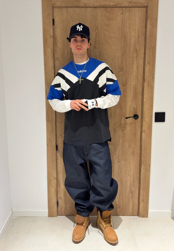 Ung man som bär en svart och blå Adidas-tröja, baggy jeans, bruna kängor, en svart New York Yankees-keps och ett korshalsband, stående inomhus.