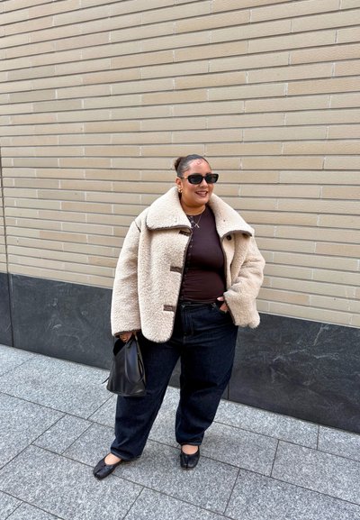 Mujer con chaqueta grande de forro de lana color beige, camiseta marrón oscuro, pantalones negros holgados, zapatos negros, gafas de sol, sosteniendo un bolso negro, de pie en la acera de la ciudad.