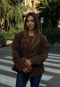 Brown suede blazer with large buttons, worn over a brown top, paired with blue jeans and a small brown handbag, on a crosswalk.