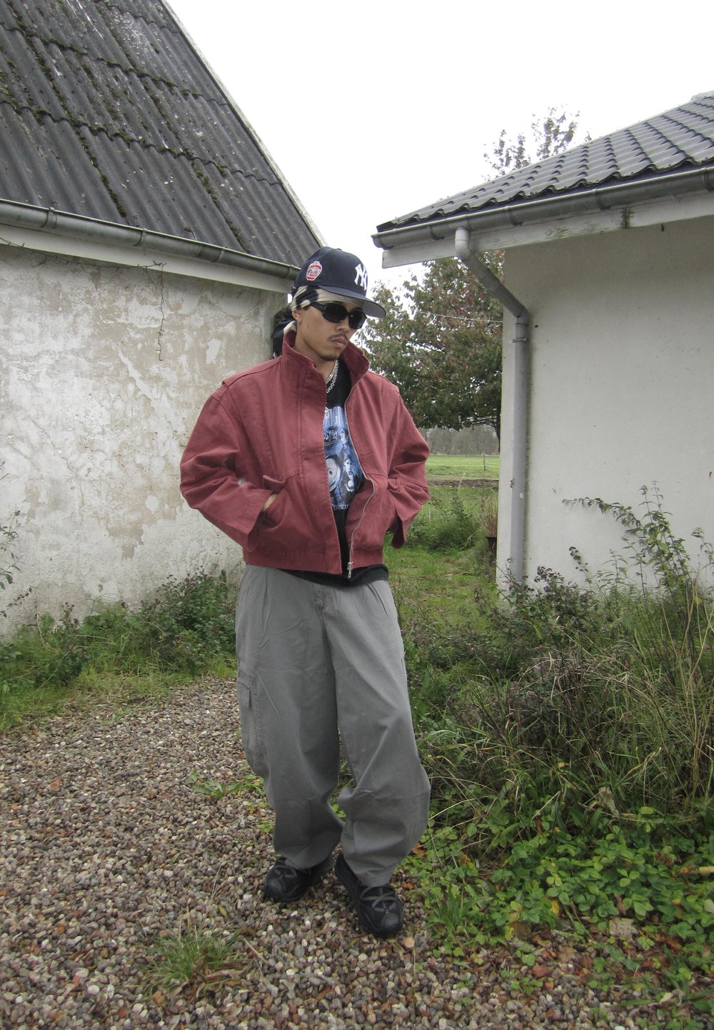 Sarkans zip-up jaku ar stāvošu apkakli, uzvilkts virs melnas T-krekli. Brīvas pelēkas kargo bikses un melni sporta apavi. Saulesbrilles un cepure noslēdz izskatu.
