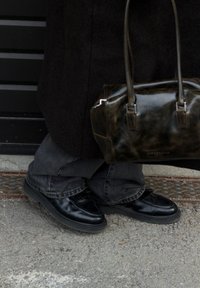 Mocassins en cuir noir avec un devant texturé, portés avec un jean gris ample. Un sac à main sombre et brillant repose à côté sur une surface grise.