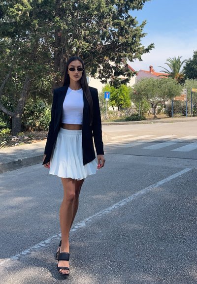 Un blazer negro, una blusa blanca corta y una falda blanca con pliegues. Lleva sandalias de plataforma negras y gafas de sol, con el cabello liso y largo.