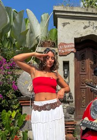 Rode geribbelde crop top, witte vloeiende rok met een decoratieve bruine riem, bloemige accenten en een gestructureerde achtergrond met groenvoorziening en een stenen bord.