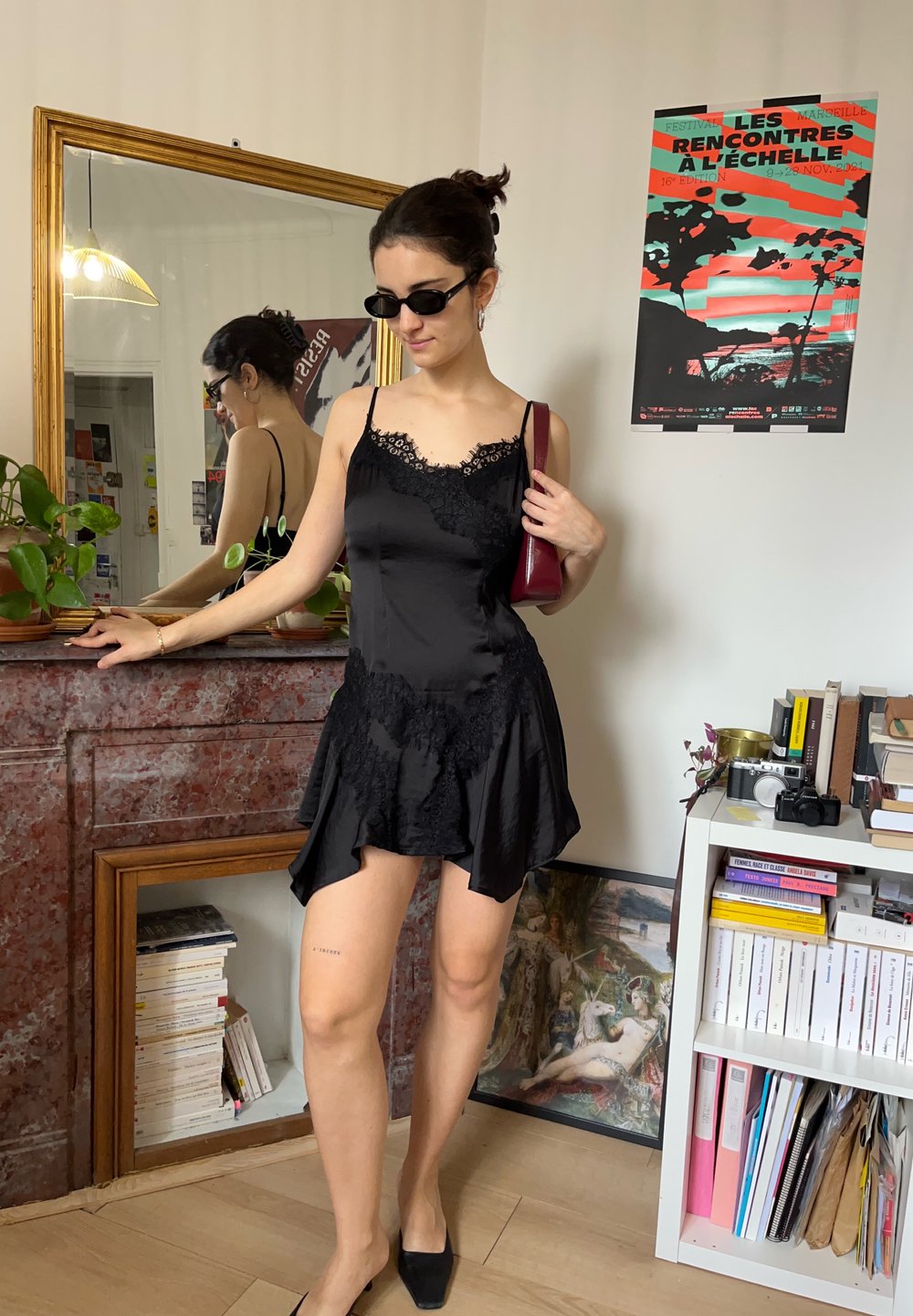 Femme en robe en dentelle noire et lunettes de soleil se tient près d'une cheminée en marbre dans une pièce avec des livres, des plantes et une affiche colorée accrochée au mur.