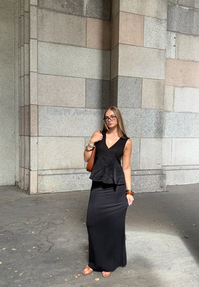 Conjunto de top peplum negro y falda larga, ambos hechos de tela suave. Los accesorios incluyen un bolso marrón y múltiples pulseras. Fondo de pared de piedra.