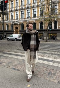 A man dressed in a dark jacket, a checkered scarf, and white trousers is walking along a cobblestone street in the city, close to a red traffic light.