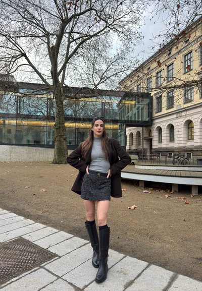 Mujer de pie con las manos en las caderas, vistiendo un suéter gris, falda negra, botas negras y un abrigo oscuro, cerca de un árbol sin hojas y un edificio histórico.