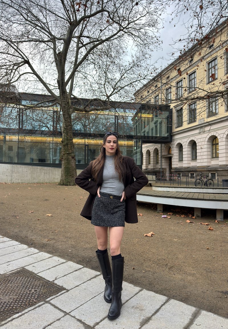 Frau steht mit Händen in der Hüfte, trägt einen grauen Pullover, schwarzen Rock, schwarze Stiefel und dunklen Mantel in der Nähe eines kahlen Baums und eines historischen Gebäudes.