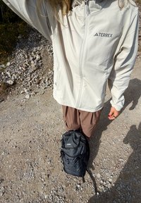 Jachetă Terrex de culoare bej deschis, cu fermoar complet, purtată cu pantaloni cargo maro. Rucsac negru pe jos, cu multiple buzunare exterioare și curele.