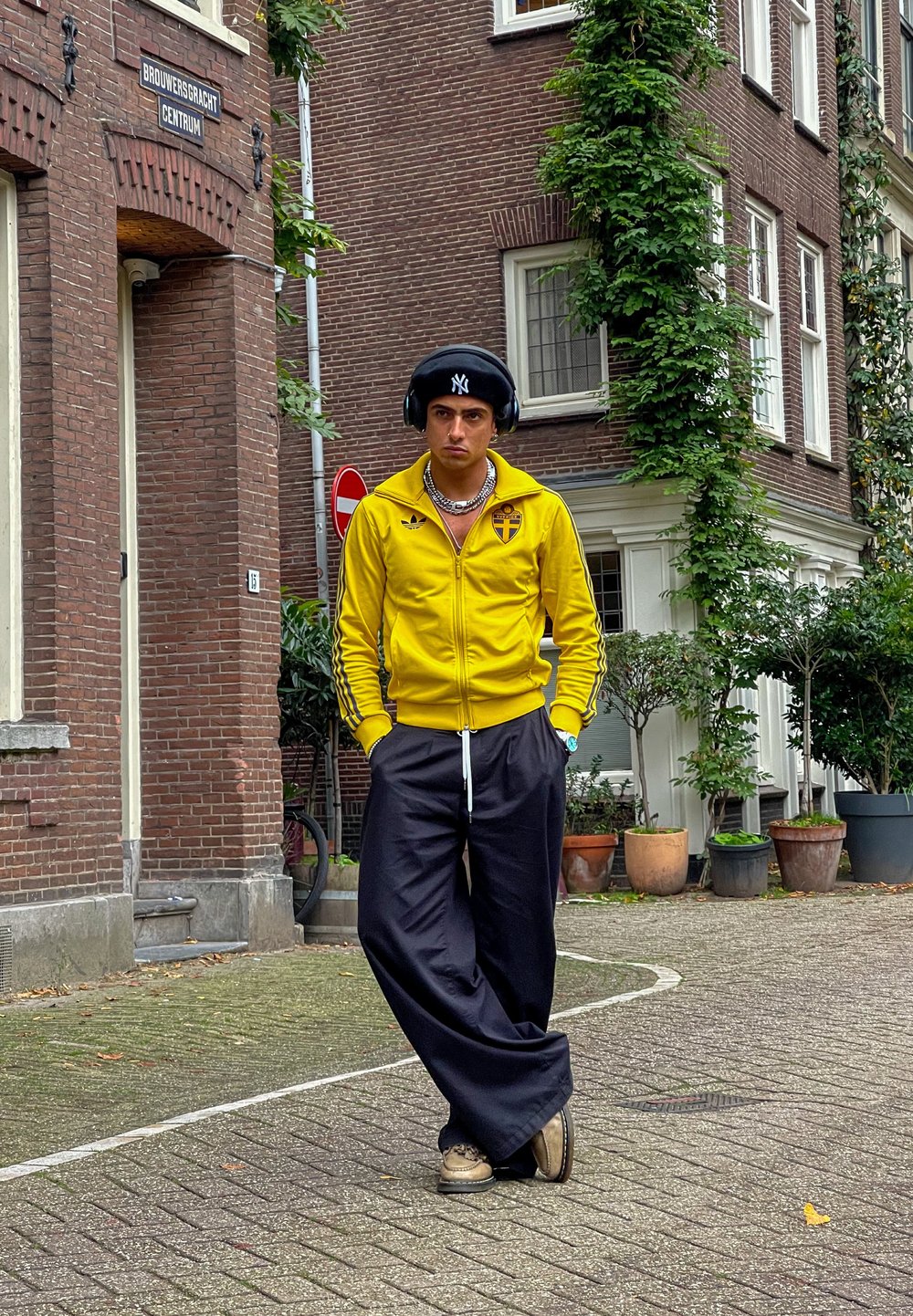 Κίτρινο μπουφάν με φερμουάρ και μαύρες ρίγες και έμβλημα, σε συνδυασμό με μαύρο φαρδύ παντελόνι. Τα αξεσουάρ περιλαμβάνουν ακουστικά και αλυσίδα στο λαιμό.
