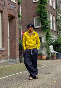 Κίτρινο μπουφάν με φερμουάρ και μαύρες ρίγες και έμβλημα, σε συνδυασμό με μαύρο φαρδύ παντελόνι. Τα αξεσουάρ περιλαμβάνουν ακουστικά και αλυσίδα στο λαιμό.