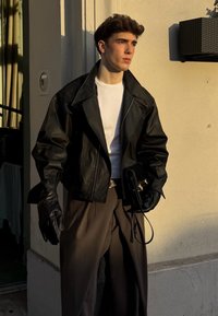 Veste en cuir noire avec larges revers, portée sur une chemise blanche. Pantalon marron, gants en cuir et un petit sac noir complètent la tenue.