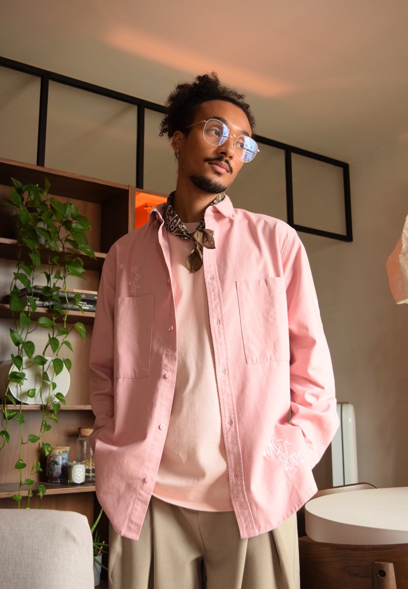 Chemise button-up rose clair avec deux poches à l'avant et broderie florale, superposée sur un T-shirt rose pâle, associée à un pantalon beige.