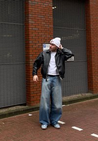 Čierna kožená bomber bunda, biele tričko, nadmerné svetlo-modré džínsy, biela čiapka a modré tenisky, stojac voči tehlovej stene.