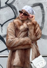 Palton din blană sintetică maro și bej cu o textura pufoasă, asortat cu o batic de mătase albă și ochelari negri de soare. Ținând o geantă albă.