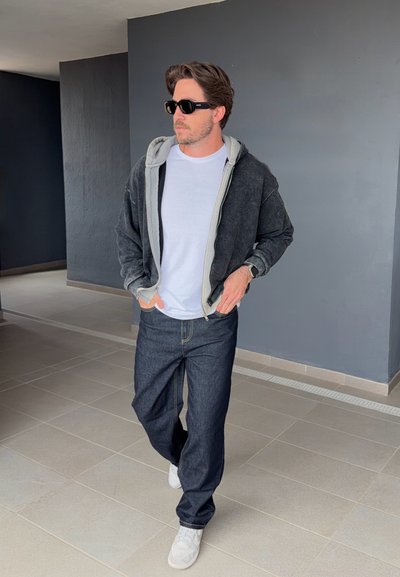 Hombre con gafas de sol, camiseta blanca, sudadera negra con cremallera, vaqueros oscuros y zapatillas blancas caminando en interiores con las manos en los bolsillos.