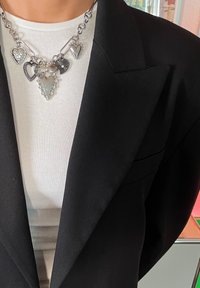 Srebrna ogrlica z različnimi obeski v obliki srca, nosena nad belim žametnim topom in črnim blazerjem, ki prikazuje plastne teksture.