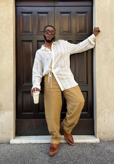 Camisa suelta a rayas crema con detalle de corbata, pantalones de lino beige y zapatos de cuero marrón. Sosteniendo un vaso transparente con una pajita. Fondo de puerta de madera.