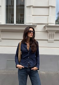 Sötétkék denim gombos ing, világosabb kék, laza szabású farmernadrággal párosítva. Barna velúrbőr válltáska, oversized sötét napszemüveg. Városi környezet.
