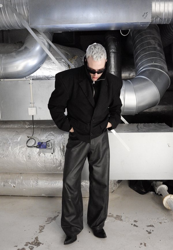 Persona con cabello corto y blanco y gafas de sol oscuras, vistiendo una chaqueta negra y pantalones anchos negros, de pie en un espacio industrial con tuberías de metal.