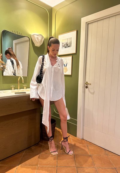 Camisa blanca de gran tamaño con mangas largas, combinada con tacones negros de tiras. Presenta una pared verde, un tocador de madera y obras de arte decorativas.