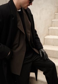 Homme portant des manteaux superposés noirs et bruns, des gants en cuir noirs et des lunettes de soleil, assis sur des escaliers en béton clair.