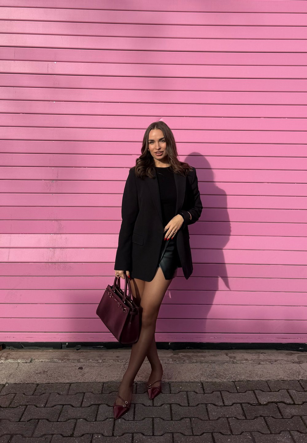 Femme en blazer noir, haut noir et short en cuir tenant un sac à main bordeaux, se tenant devant un mur à panneaux horizontaux roses.