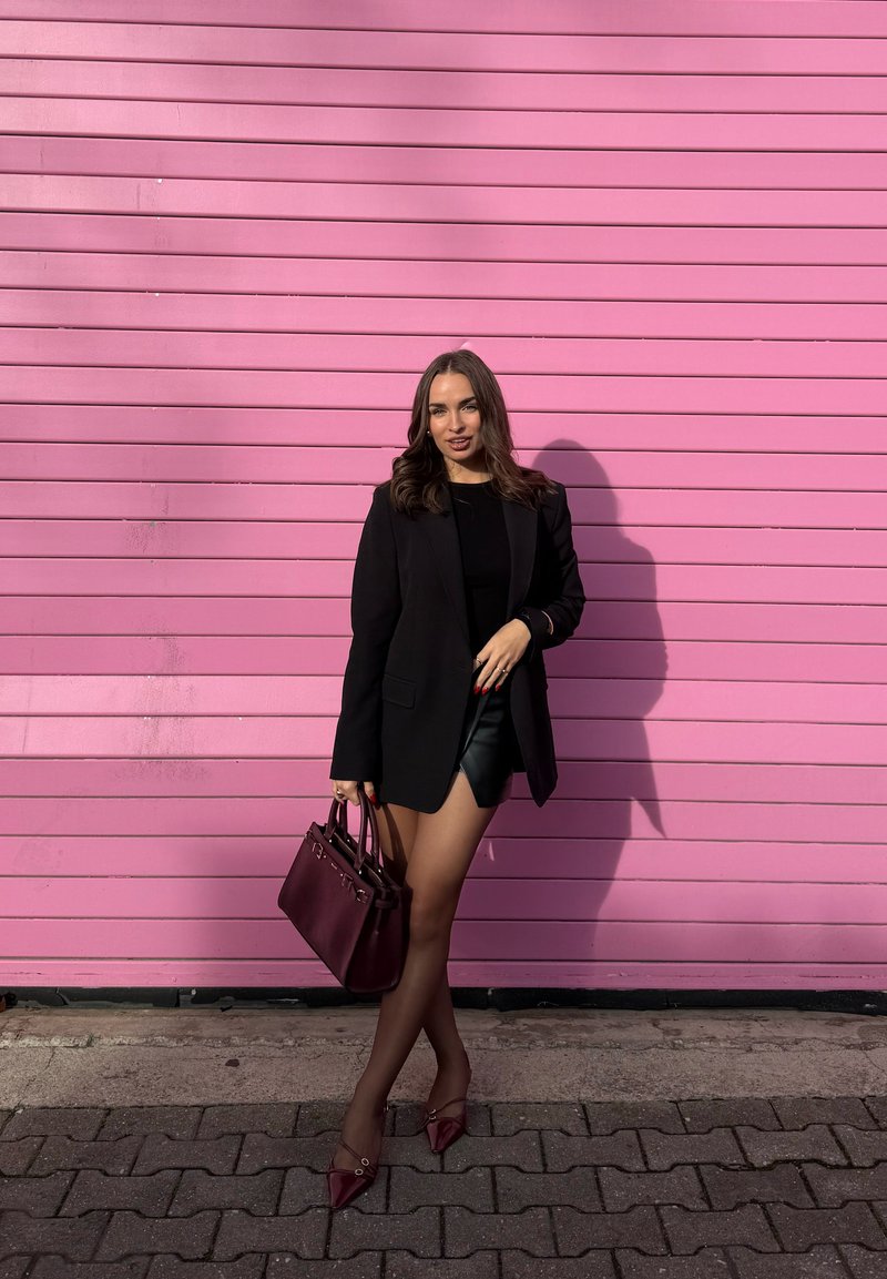Femme en blazer noir, haut noir et short en cuir tenant un sac à main bordeaux, se tenant devant un mur à panneaux horizontaux roses.