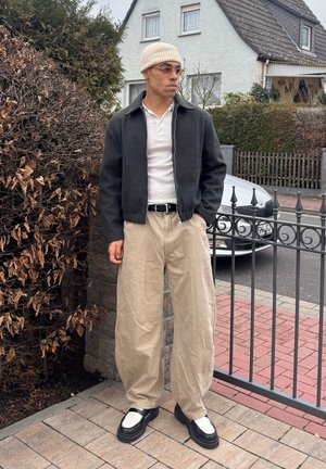 Joven con gorro beige, gafas tintadas, chaqueta oscura, camisa blanca, pantalones anchos beige y zapatos blanco y negro, de pie cerca de una cerca residencial.