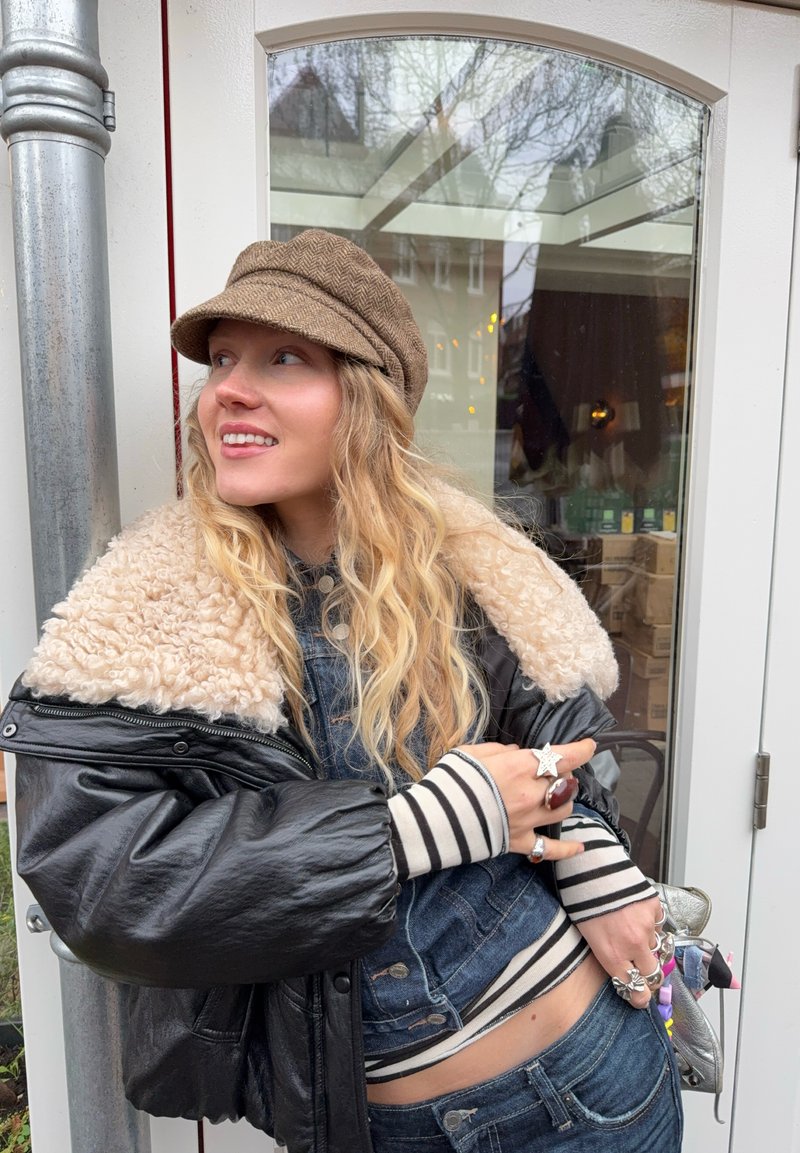 Junge Frau mit langen blonden Haaren trägt braune Mütze, Lederjacke mit Schaffellkragen, gestreiftes Shirt und Ringe, lehnt an einer Glastür.