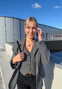 Šedý kostkovaný blazér cez šedú vestu s gombíkmi, doplnený čiernymi nohavicami. Kruhový prívesok a taška na rameno. Vonkajšie prostredie s modrou oblohou.