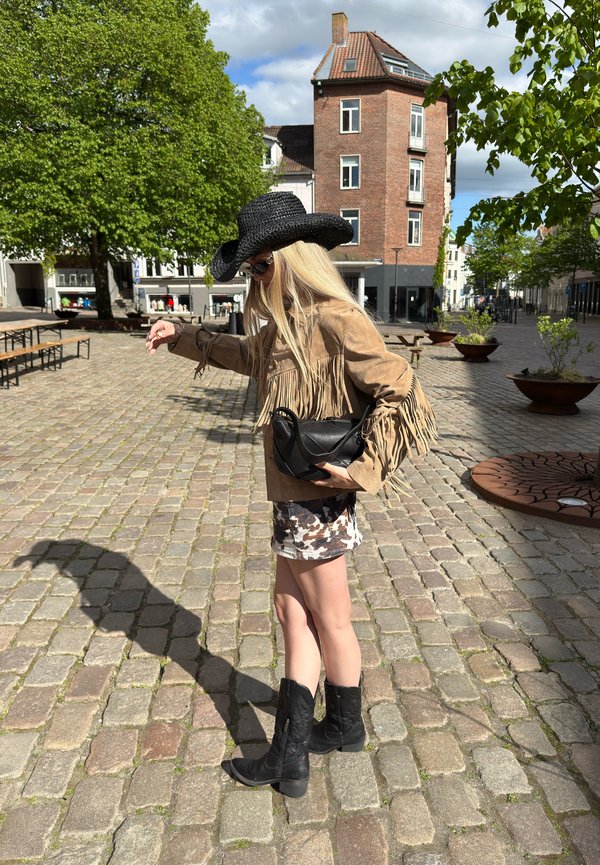 Osoba nosící černý kovbojský klobouk, třásňovanou hnědou bundu, černé boty a vzorovanou sukni stojí na dlážděné ulici ve slunné městské oblasti.