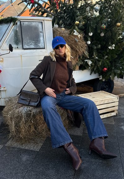 Jersey de cuello alto marrón, abrigo gris, gorra azul y gafas de sol; jeans acampanados y botines marrón oscuro. Sentada sobre pacas de heno cerca del árbol de Navidad.