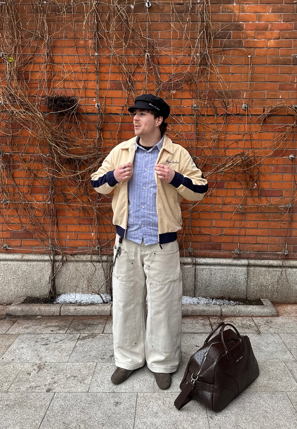 Jeune homme portant une veste beige, une chemise bleu clair, un pantalon large blanc et une casquette noire, debout près d'un mur de briques couvert de vignes sans feuilles avec un sac en cuir marron posé au sol.