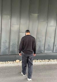 Crni vuneni pulover, crne cargo hlače i bijele tenisice. Stoji naslonjen na glatku sivu zid s naglaskom na kamenčiće u donjem dijelu.